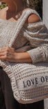 A woman wearing a cream crochet knit off-the-shoulder top and dark pants carries a large cream crochet tote bag with black text reading 'LOVE OF IVY'.