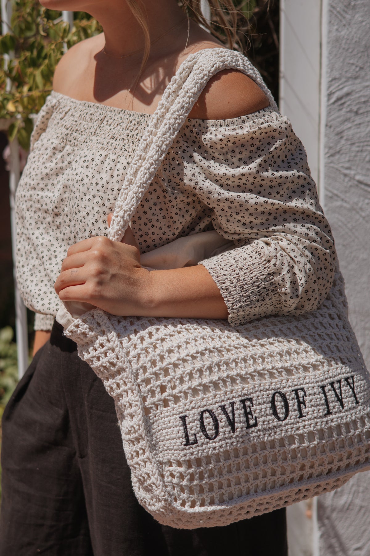 A woman wearing a cream crochet knit off-the-shoulder top and dark pants carries a large cream crochet tote bag with black text reading 'LOVE OF IVY'.