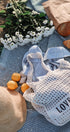White mesh bag with 'LOVE OF' text on a blue blanket with flowers, oranges, and a hat.