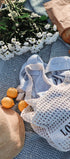 White mesh bag with 'LOVE OF' text on a blue blanket with flowers, oranges, and a hat.
