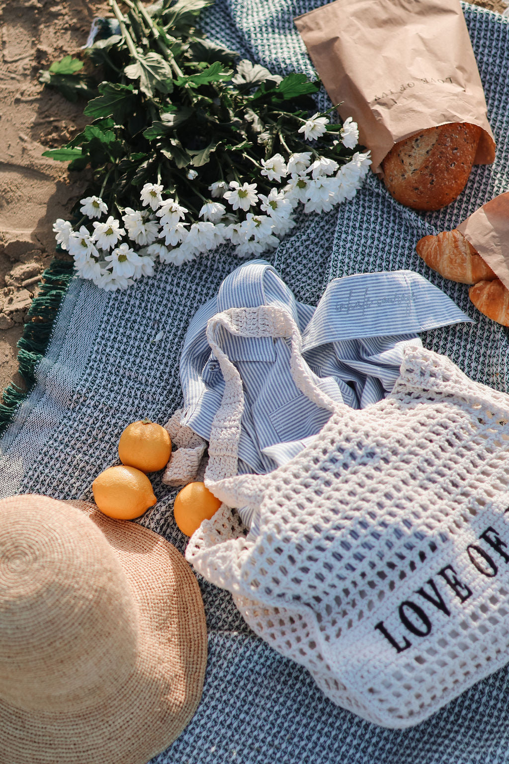 White mesh bag with 'LOVE OF' text on a blue blanket with flowers, oranges, and a hat.