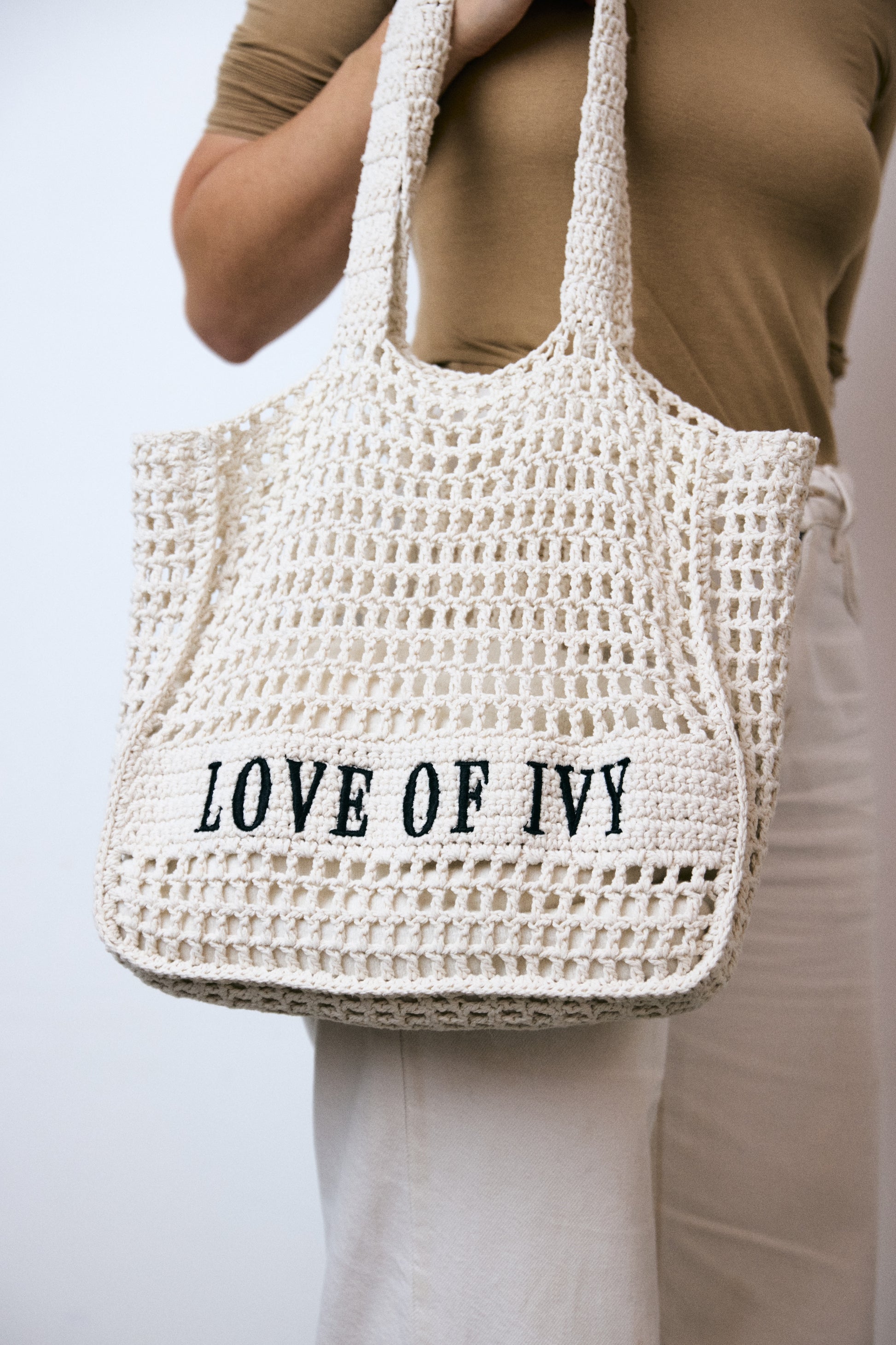 White woven bag with 'LOVE OF IVY' text held by a person wearing a beige top and white pants.
