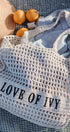 Mesh bag with 'LOVE OF IVY' text on a textured surface with oranges and a hat.