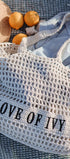 Mesh bag with 'LOVE OF IVY' text on a textured surface with oranges and a hat.
