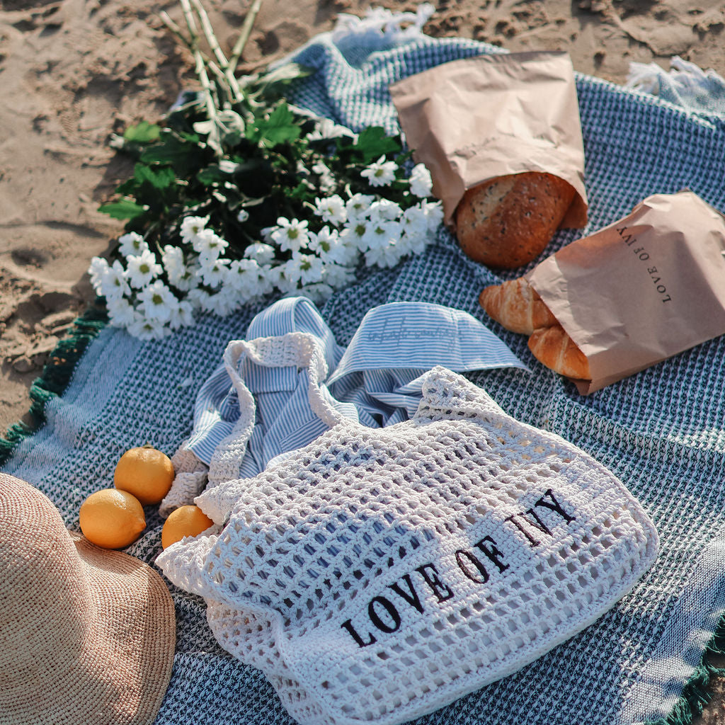 Picnic setup on a sandy beach with a blue blanket, bag labeled 'LOVE OF IT', bread, oranges, and flowers.