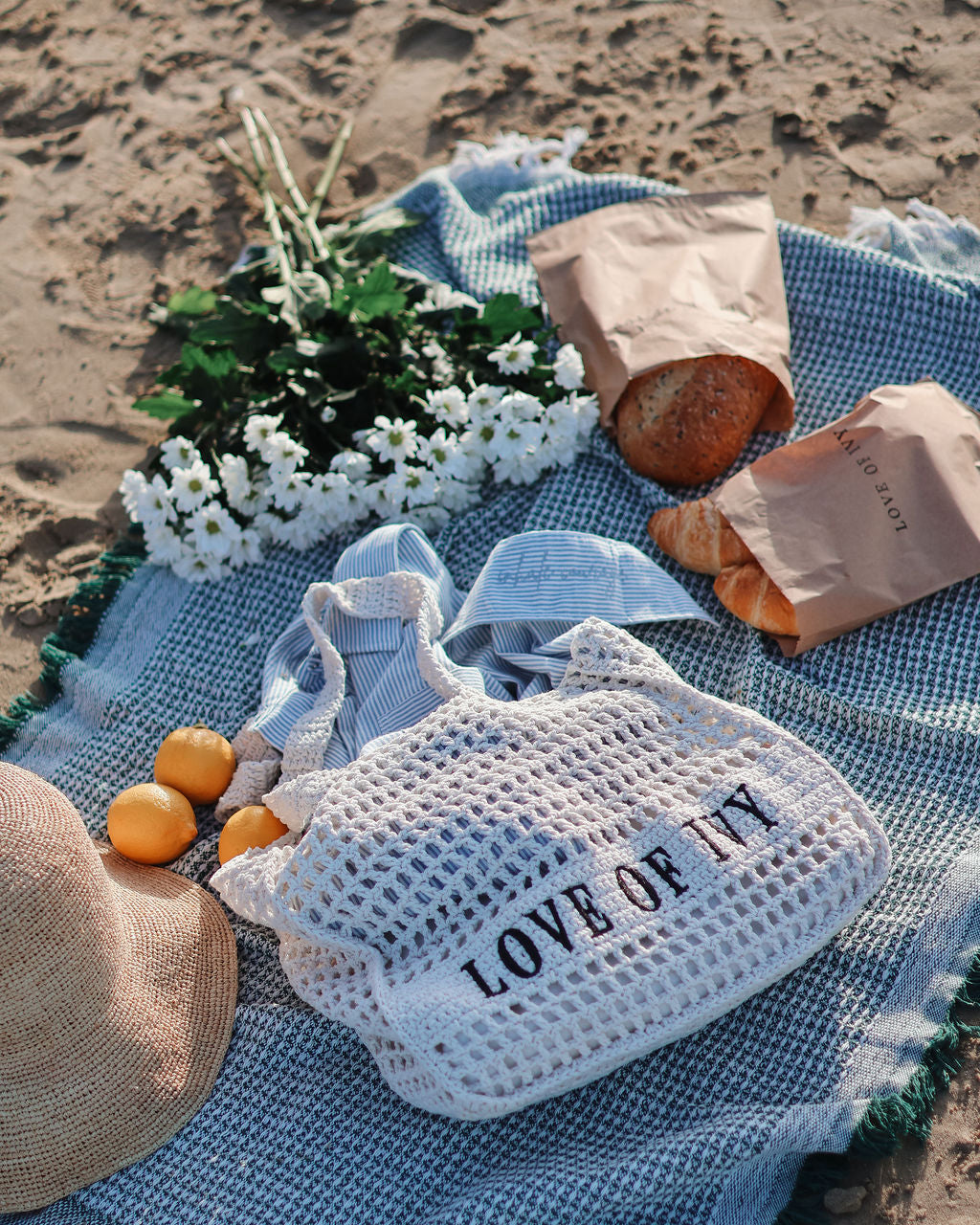 Picnic setup on a sandy beach with a blue blanket, bag labeled 'LOVE OF IT', bread, oranges, and flowers.