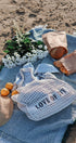 Picnic setup on a sandy beach with a blue blanket, bag labeled 'LOVE OF IT', bread, oranges, and flowers.