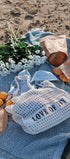 Picnic setup on a sandy beach with a blue blanket, bag labeled 'LOVE OF IT', bread, oranges, and flowers.