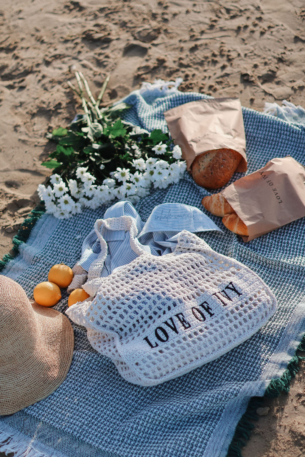 Picnic setup on a sandy beach with a blue blanket, bag labeled 'LOVE OF IT', bread, oranges, and flowers.