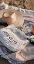 Woven bag with 'LOVE OF IVY' text on a beach setting with a hat, sunglasses, and orange.