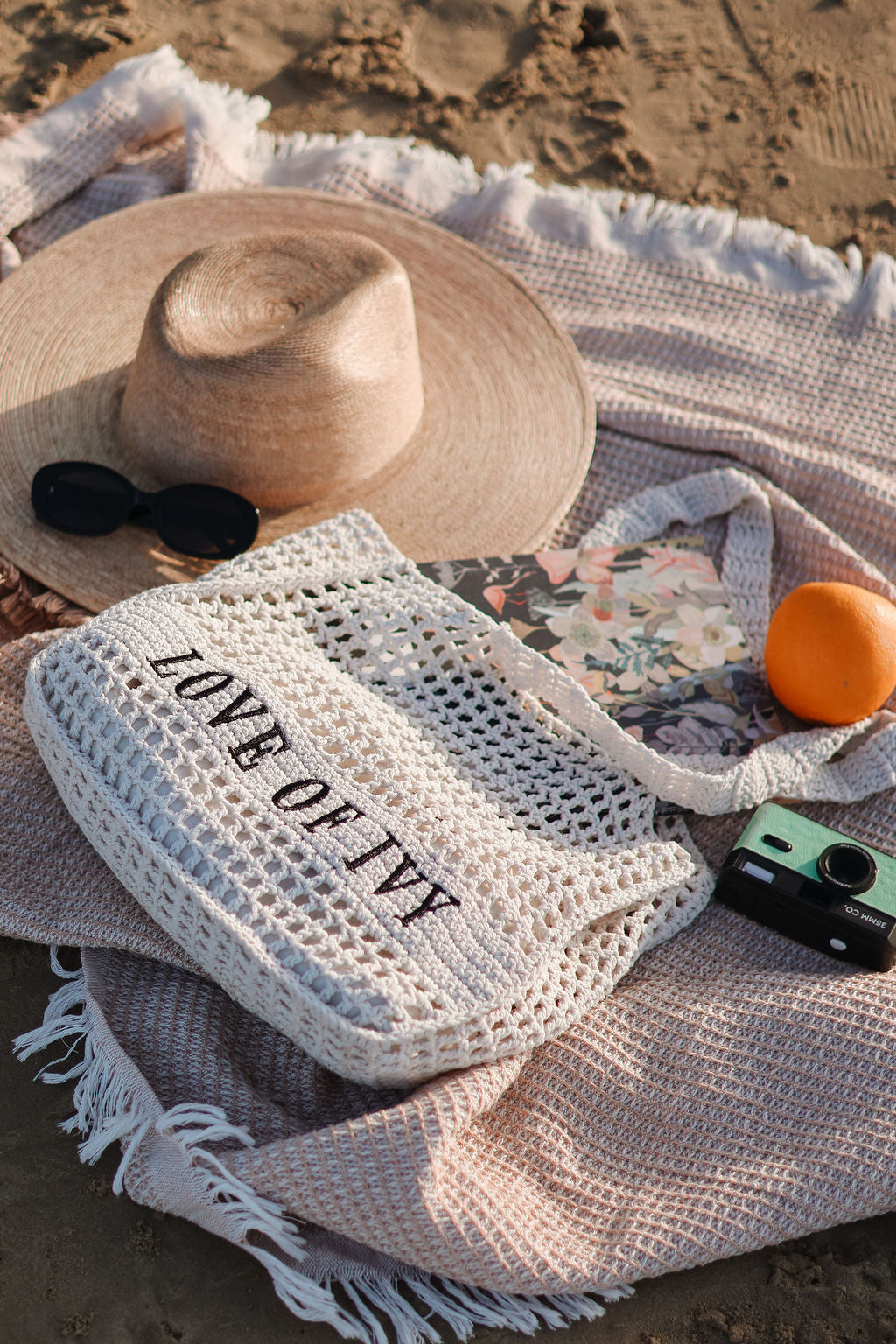 Woven bag with 'LOVE OF IVY' text on a beach setting with a hat, sunglasses, and orange.