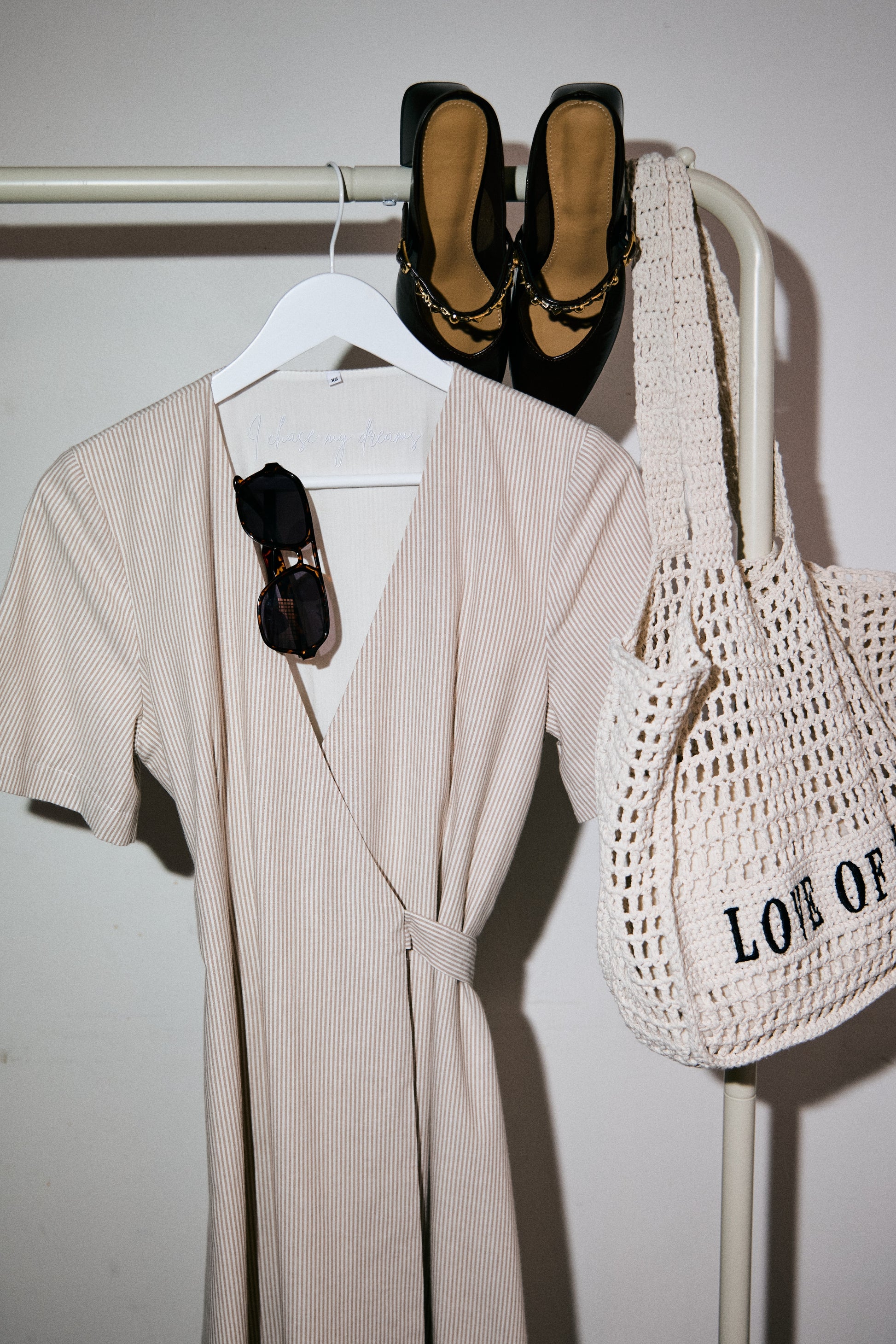 Beige dress, black shoes, and a white mesh bag on a rack against a white wall.