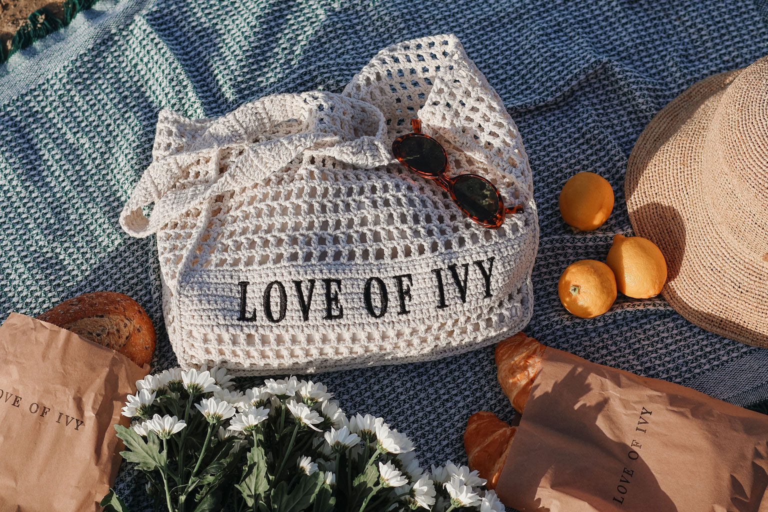 White crochet bag with 'LOVE OF IVY' text on a blue textured surface with oranges and a hat.