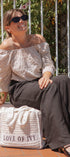 A woman wearing a cream off-the-shoulder top with a small black floral pattern and dark wide-leg trousers sits outdoors on a brick patio. She is holding a large white crochet tote bag with the text 'LOVE OF IVY' printed on it.