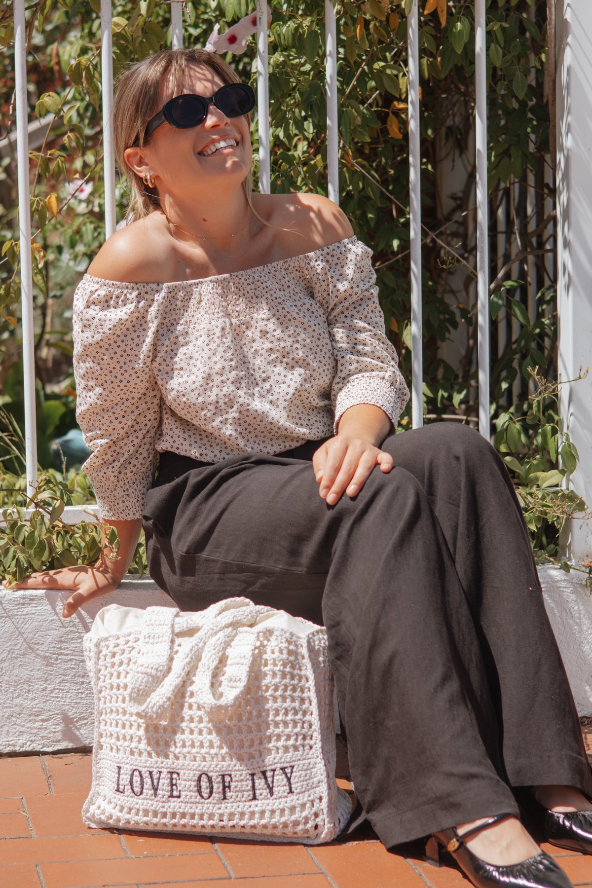 A woman wearing a cream off-the-shoulder top with a small black floral pattern and dark wide-leg trousers sits outdoors on a brick patio. She is holding a large white crochet tote bag with the text 'LOVE OF IVY' printed on it.