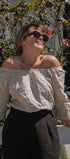 A woman outdoors wearing an off-the-shoulder long-sleeve blouse with a small beige and white polka dot pattern, paired with black trousers and dark sunglasses.