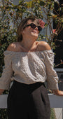 A woman outdoors wearing an off-the-shoulder long-sleeve blouse with a small beige and white polka dot pattern, paired with black trousers and dark sunglasses.