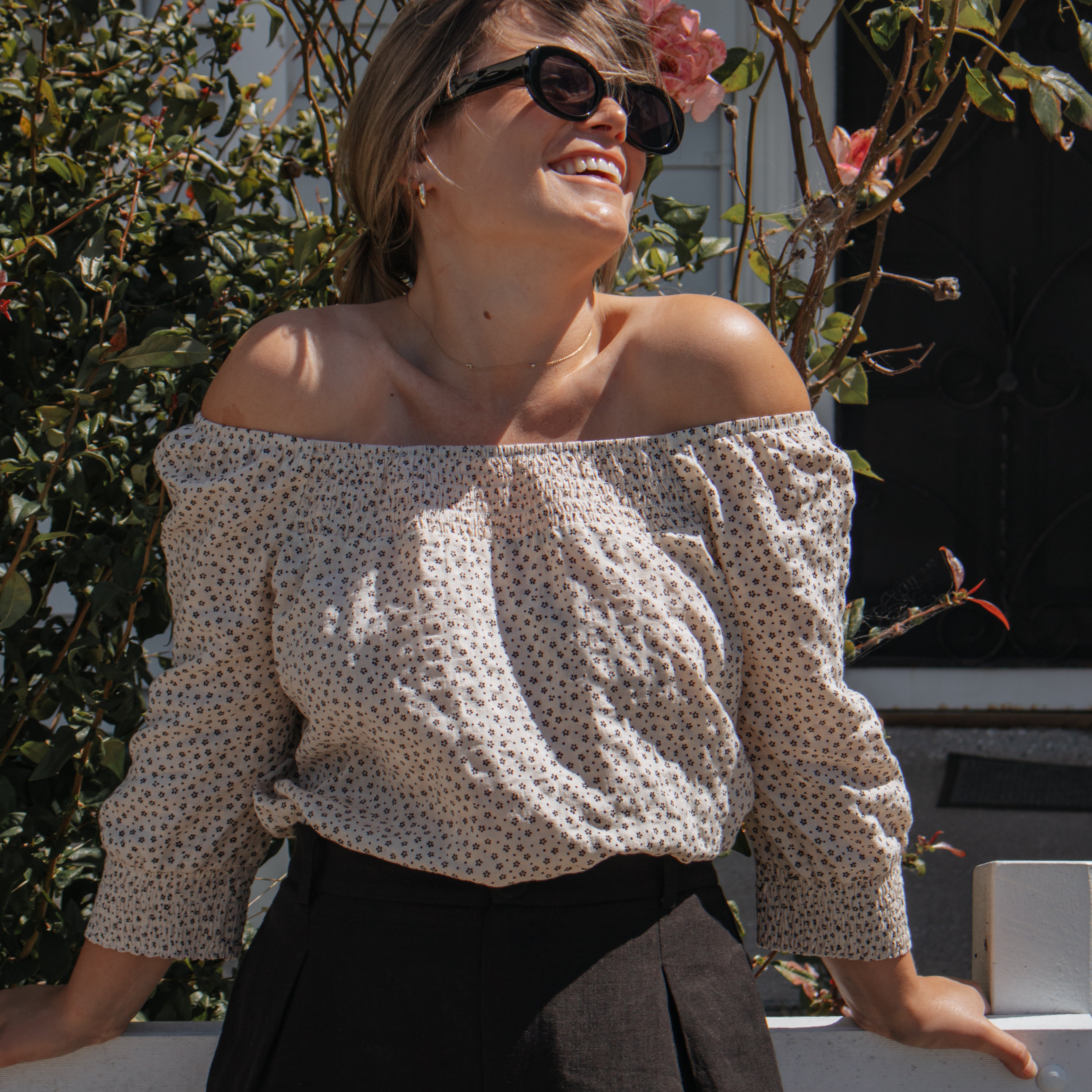 A woman outdoors wearing an off-the-shoulder long-sleeve blouse with a small beige and white polka dot pattern, paired with black trousers and dark sunglasses.