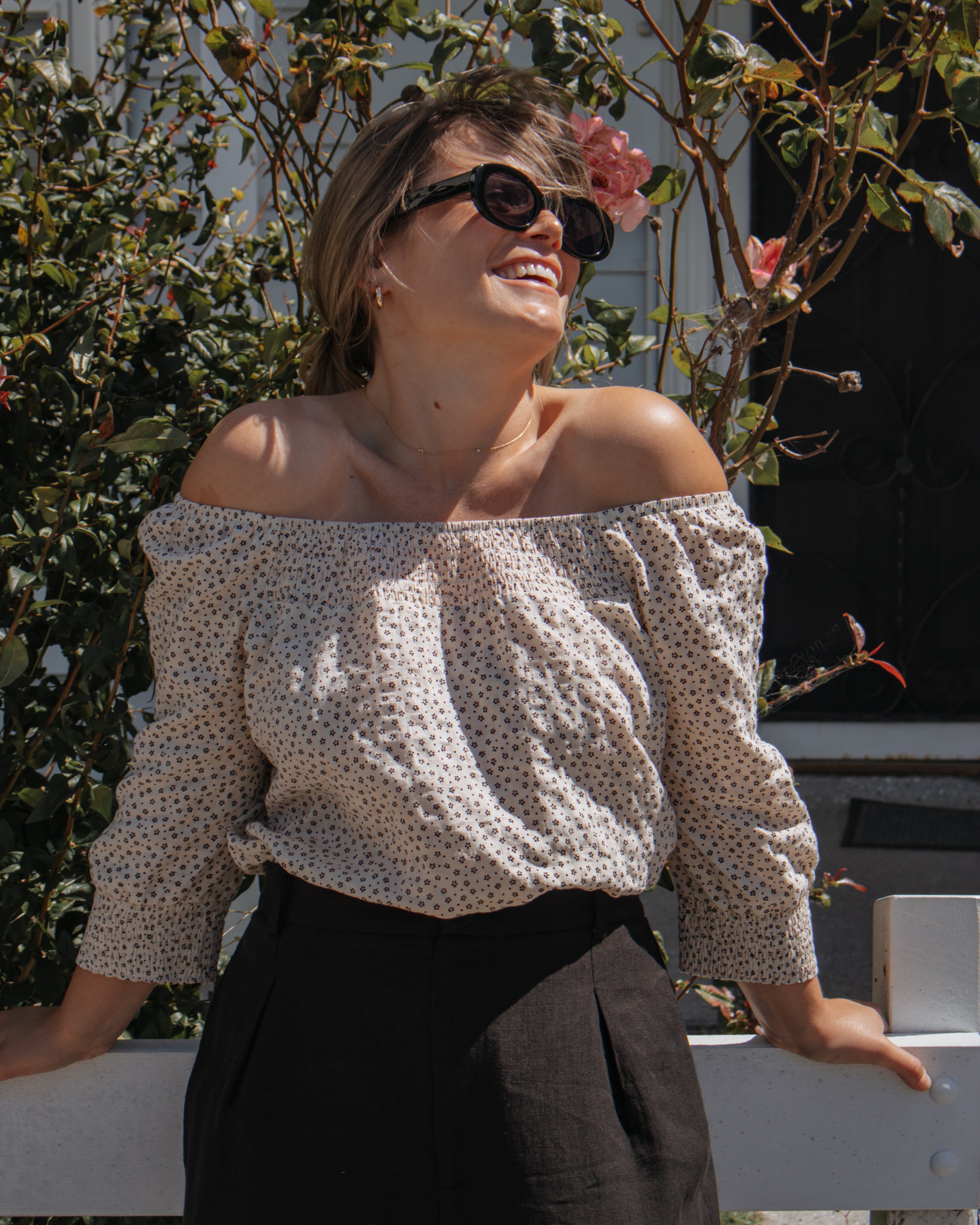 A woman outdoors wearing an off-the-shoulder long-sleeve blouse with a small beige and white polka dot pattern, paired with black trousers and dark sunglasses.