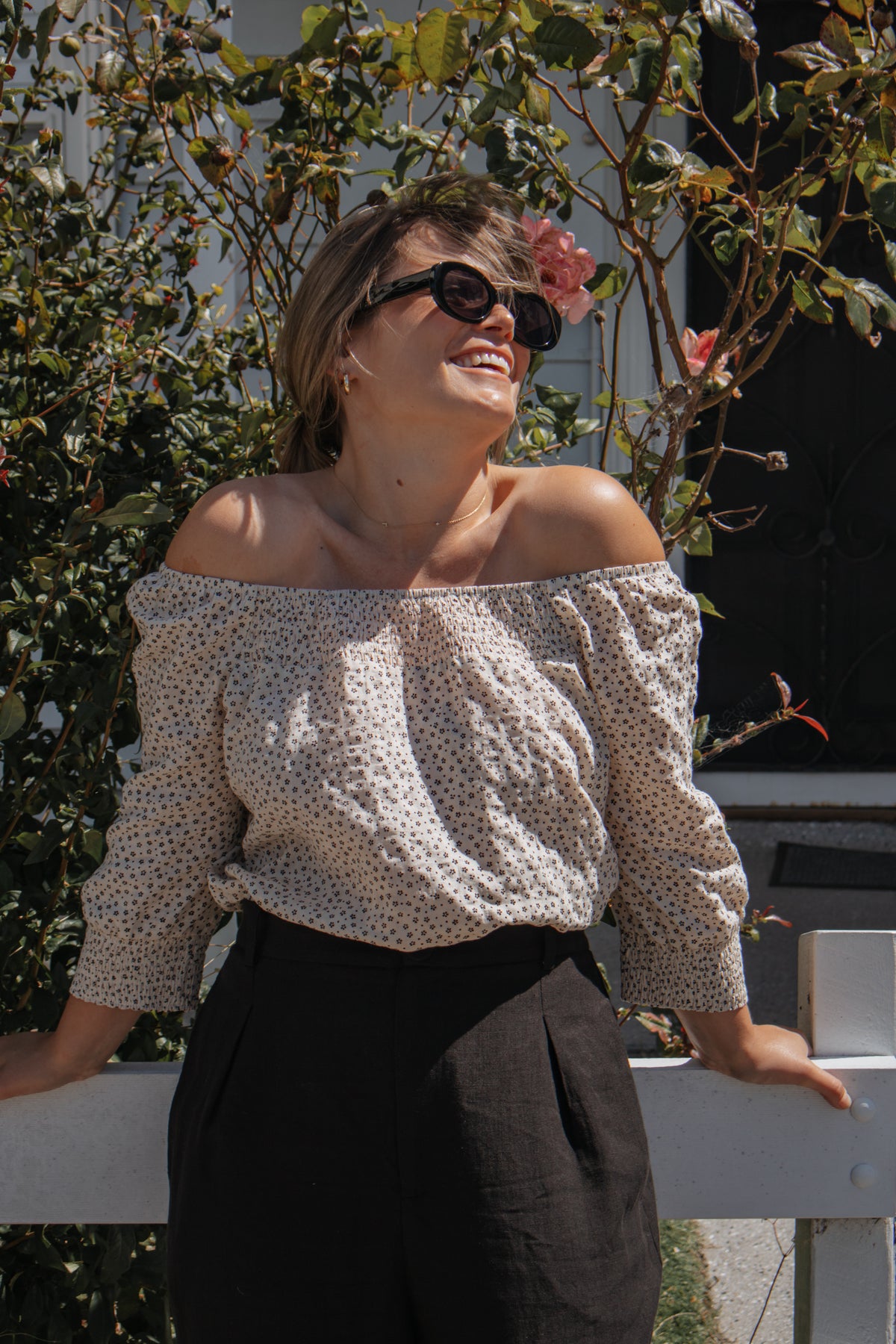 A woman outdoors wearing an off-the-shoulder long-sleeve blouse with a small beige and white polka dot pattern, paired with black trousers and dark sunglasses.