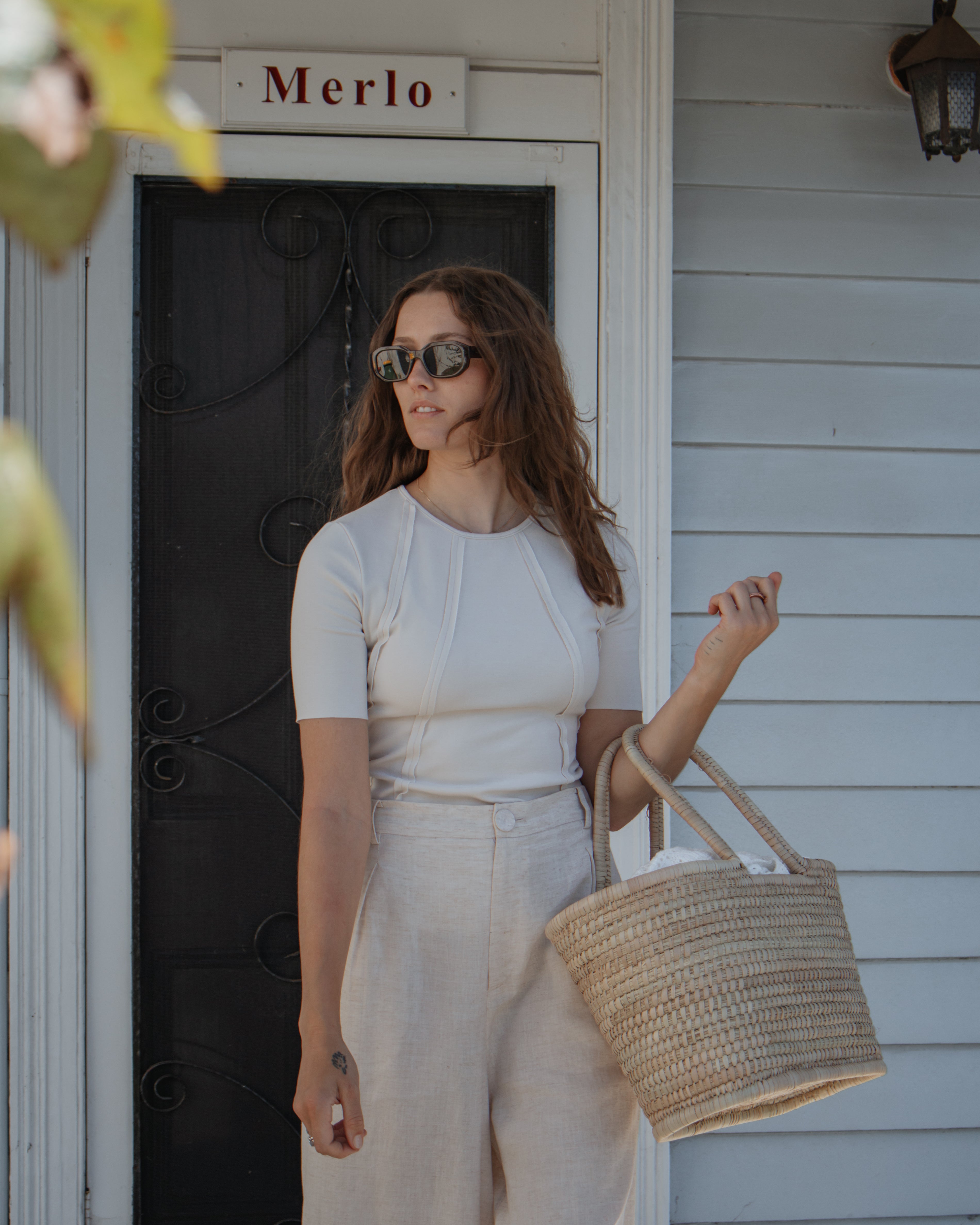 A woman wearing a white short-sleeve top and cream wide-leg pants stands on a porch holding a woven straw tote bag. The door behind her has a sign reading 'Merlo'.