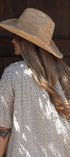 A woman shown from the back wearing a wide-brimmed straw hat and a cream-colored short-sleeve dress with a small dark floral pattern, standing against a dark wooden door.