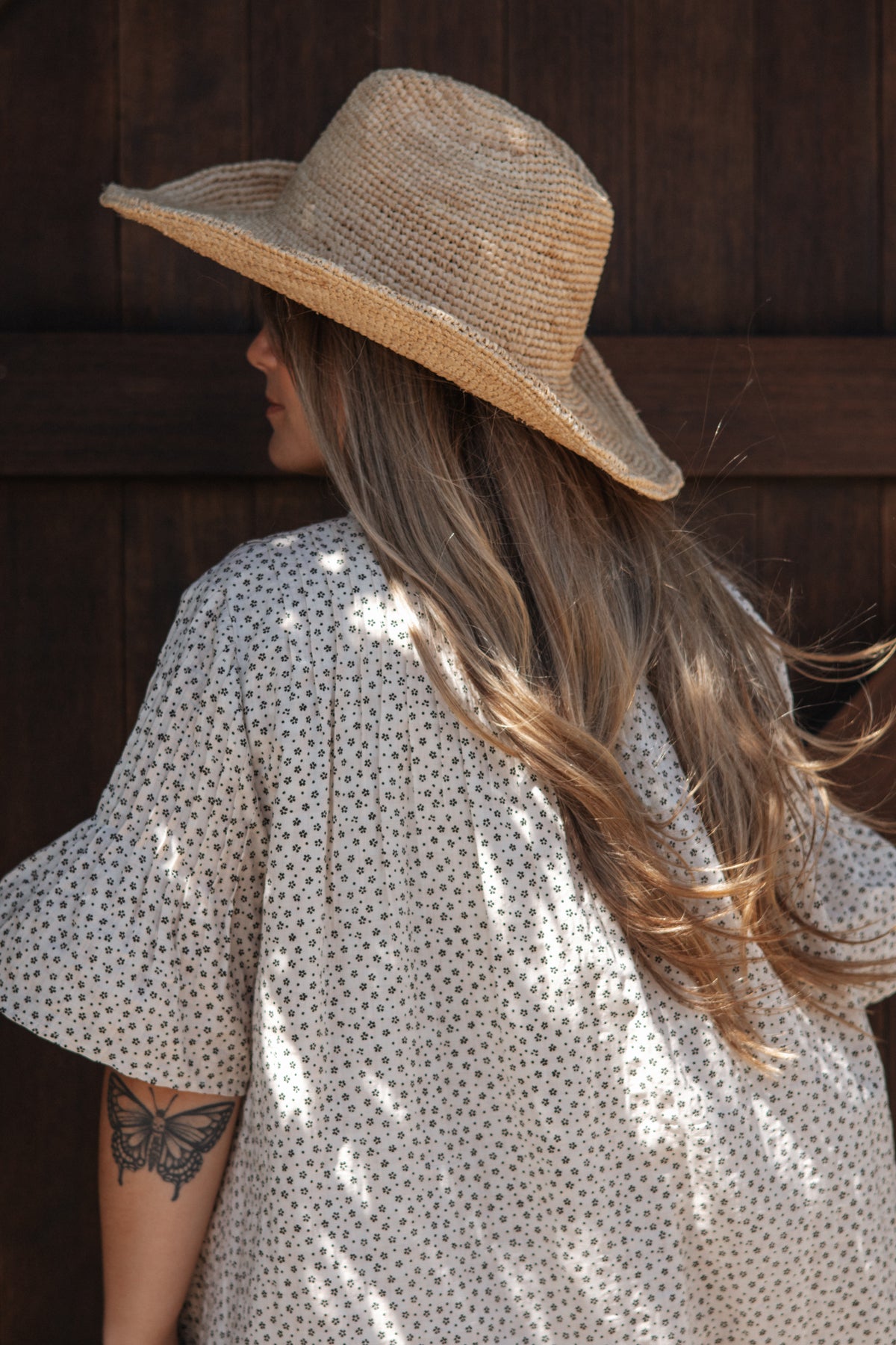 A woman shown from the back wearing a wide-brimmed straw hat and a cream-colored short-sleeve dress with a small dark floral pattern, standing against a dark wooden door.