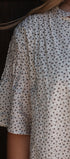 A close-up of a person wearing a loose white blouse with a small repeating brown floral or dot pattern and a tie detail at the neckline.