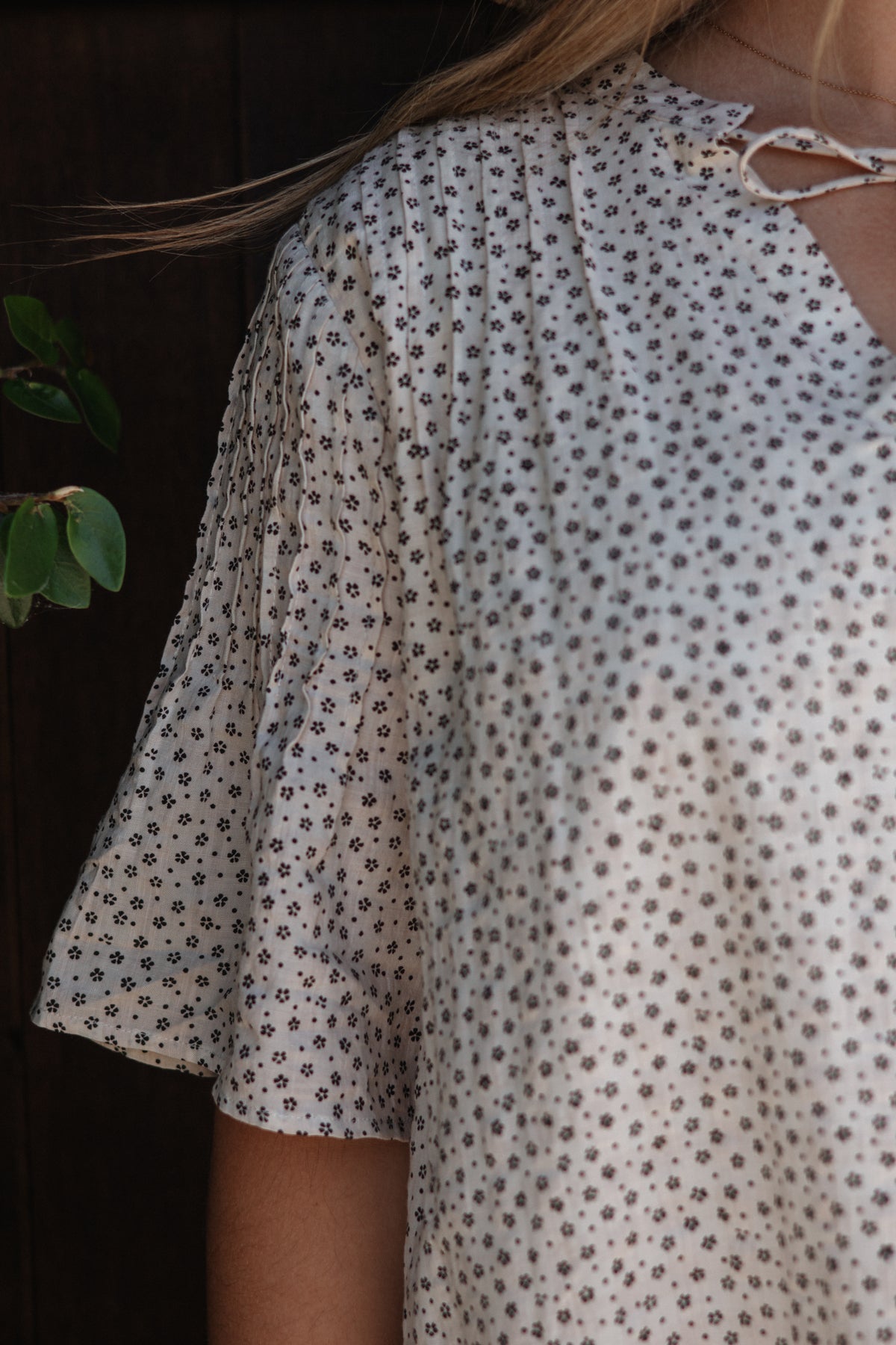 A close-up of a person wearing a loose white blouse with a small repeating brown floral or dot pattern and a tie detail at the neckline.