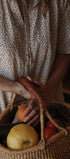 A person wearing a cream-colored short-sleeve dress with a small brown polka dot pattern holds a woven basket filled with fresh vegetables and fruit, including a carrot, zucchini, tomato, and pear.