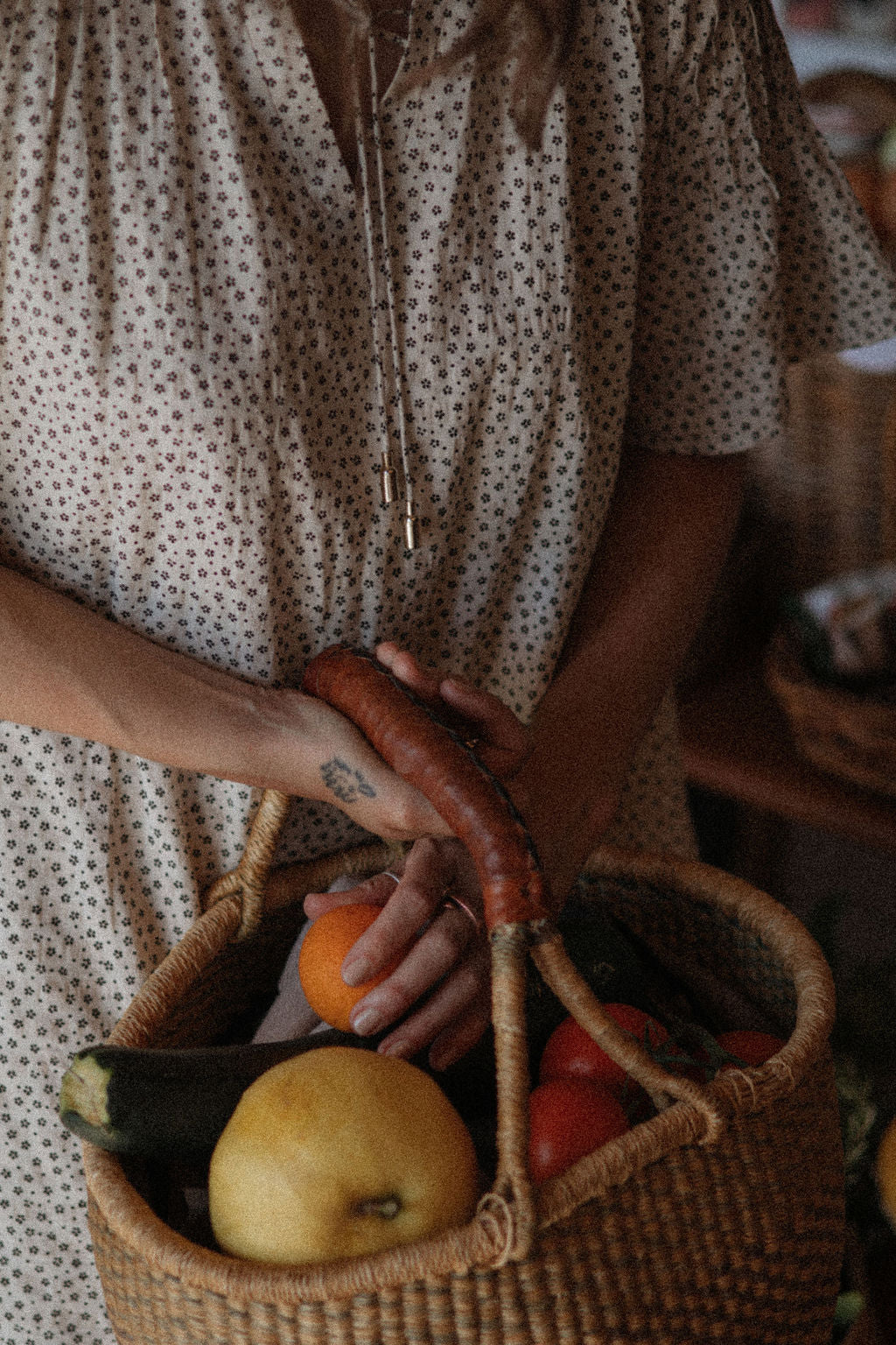 A person wearing a cream-colored short-sleeve dress with a small brown polka dot pattern holds a woven basket filled with fresh vegetables and fruit, including a carrot, zucchini, tomato, and pear.