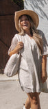 A woman laughing outdoors while wearing a cream short-sleeve mini dress with a small dark floral print, a wide-brim straw hat, and carrying a matching woven tote bag.