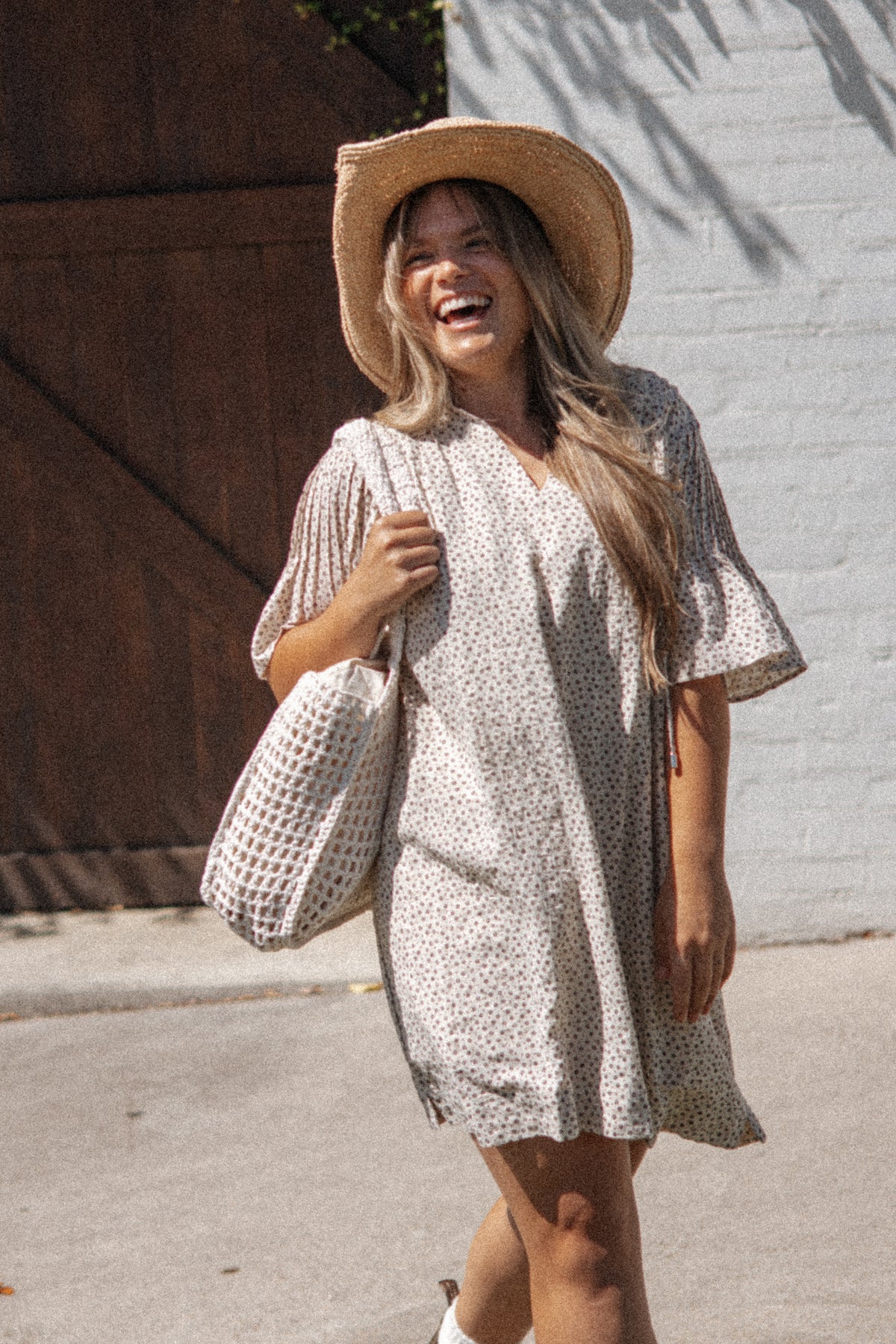 A woman laughing outdoors while wearing a cream short-sleeve mini dress with a small dark floral print, a wide-brim straw hat, and carrying a matching woven tote bag.