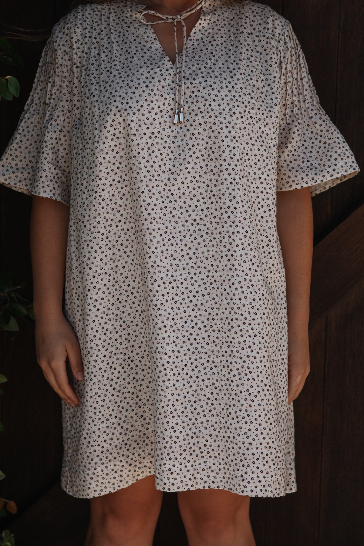 A person wearing a loose short-sleeve tunic dress with a small dark floral print on a light cream background, featuring a tie-neck detail.