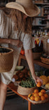 A woman wearing a straw hat and a cream-colored short-sleeve dress with a small white pattern reaches for an orange on a wooden table filled with fresh produce and woven baskets in a rustic shop setting.