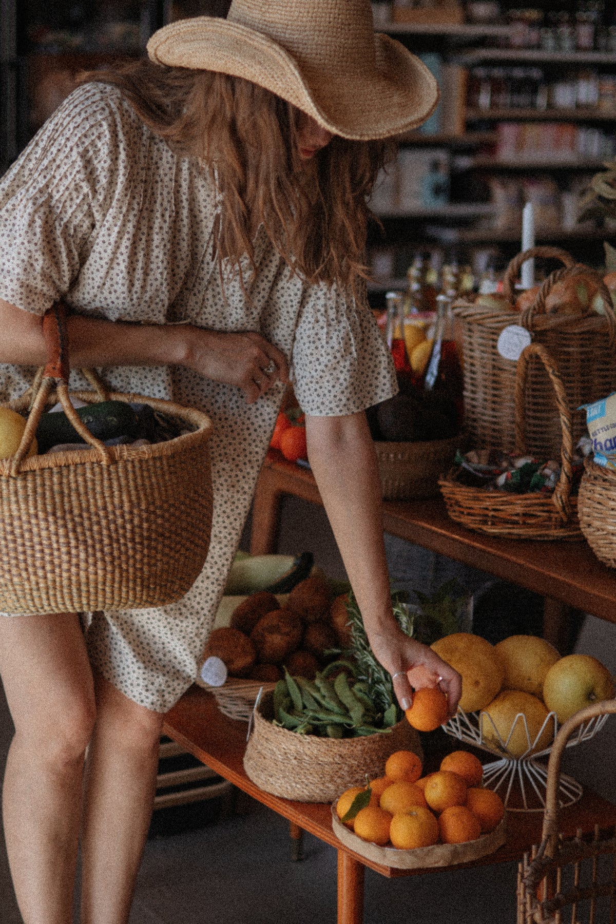 A woman wearing a straw hat and a cream-colored short-sleeve dress with a small white pattern reaches for an orange on a wooden table filled with fresh produce and woven baskets in a rustic shop setting.