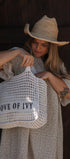 A woman wearing a straw cowboy hat and a cream polka-dot dress holds a large white crochet totebeach  bag with the text 'LOVE OF IVY' printed on the front, set against a dark wooden background.