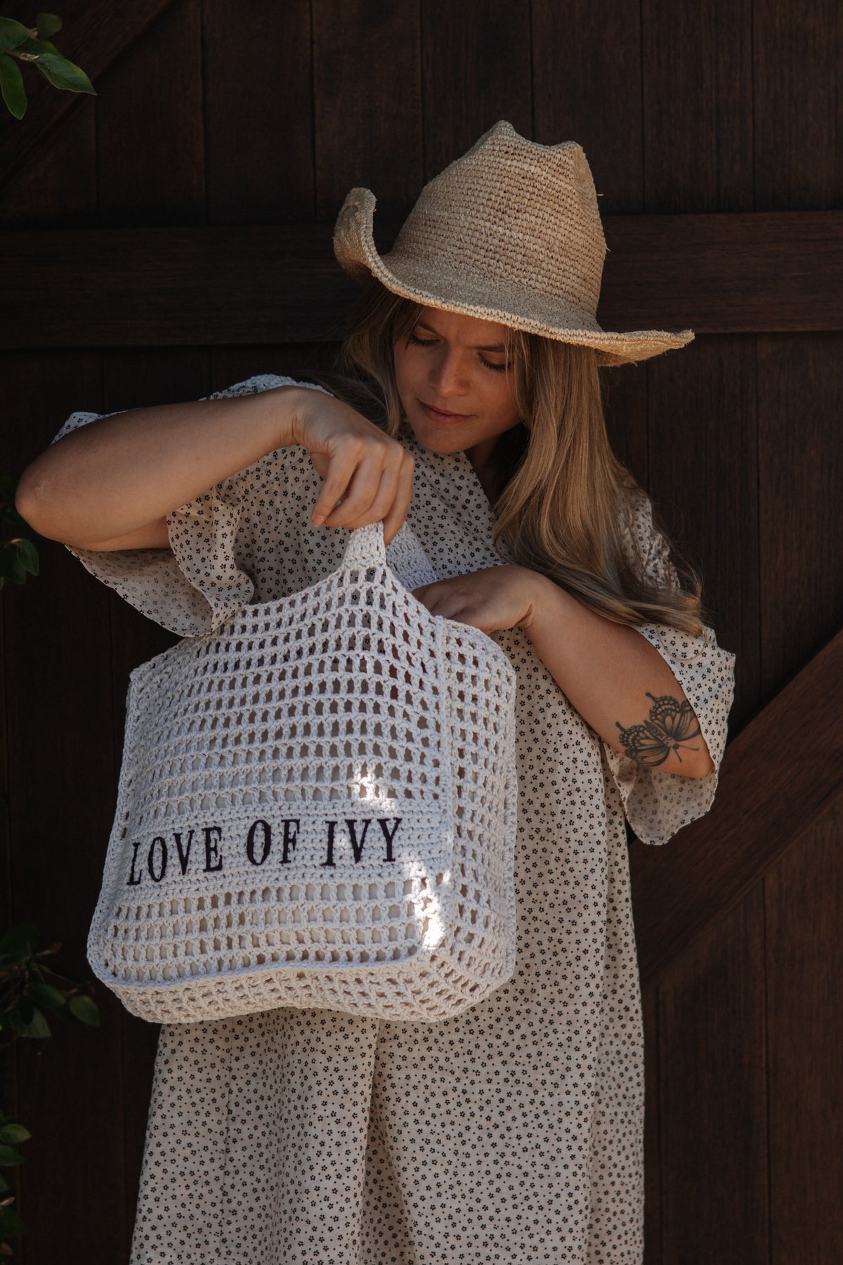 A woman wearing a straw cowboy hat and a cream polka-dot dress holds a large white crochet totebeach  bag with the text 'LOVE OF IVY' printed on the front, set against a dark wooden background.
