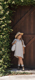 A woman stands in front of a large dark brown wooden barn-style door, wearing a light beige patterned dress, a wide-brim straw hat, and carrying a white woven shoulder bag.