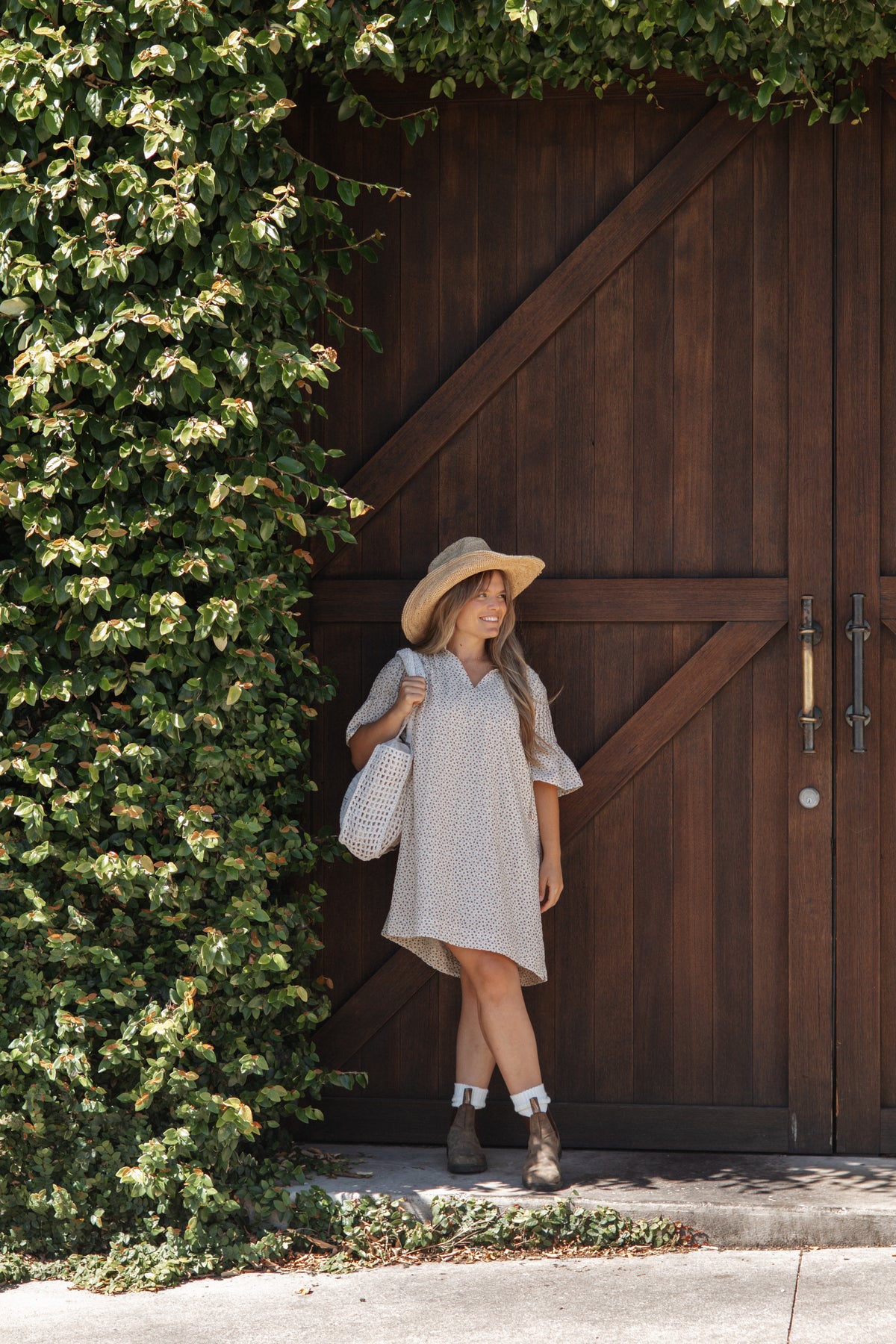A woman stands in front of a large dark brown wooden barn-style door, wearing a light beige patterned dress, a wide-brim straw hat, and carrying a white woven shoulder bag.