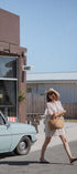 A woman in a white dress and straw hat walks across a street carrying a woven straw tote bag, with a vintage car and a building labeled BONES in the background.