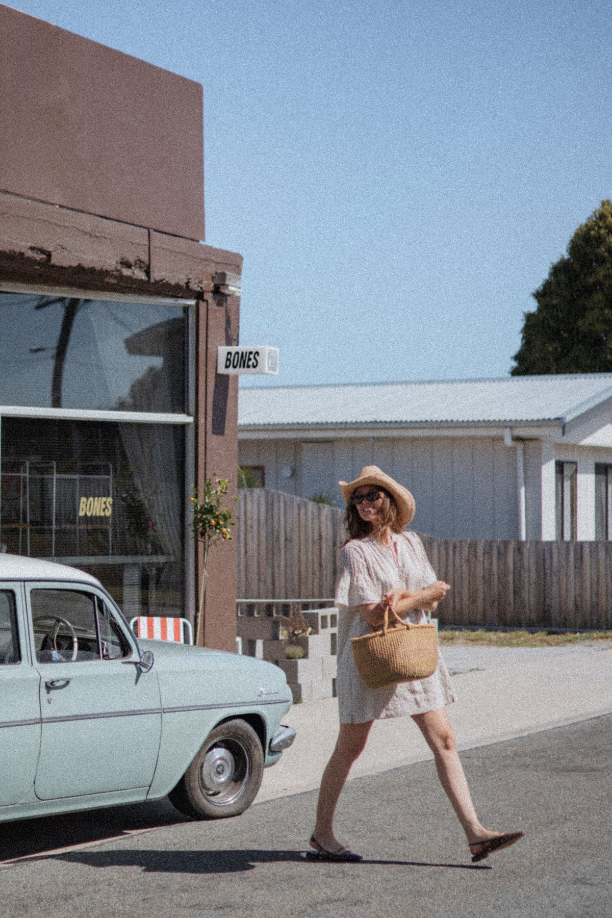 A woman in a white dress and straw hat walks across a street carrying a woven straw tote bag, with a vintage car and a building labeled BONES in the background.