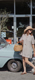 A woman wearing a beige short-sleeve button-front dress, a straw hat, and sunglasses walks past a vintage light blue car in front of a storefront.