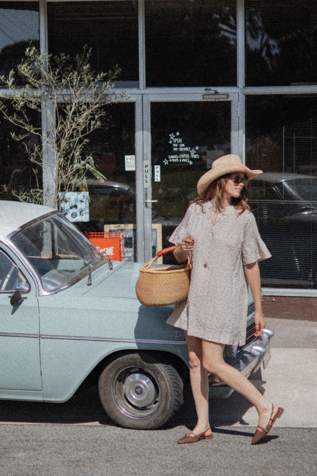 A woman wearing a beige short-sleeve button-front dress, a straw hat, and sunglasses walks past a vintage light blue car in front of a storefront.