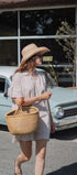 A woman walking outdoors in front of a vintage car and storefront, wearing a light beige patterned dress, wide-brim straw hat, and carrying a woven straw tote bag.