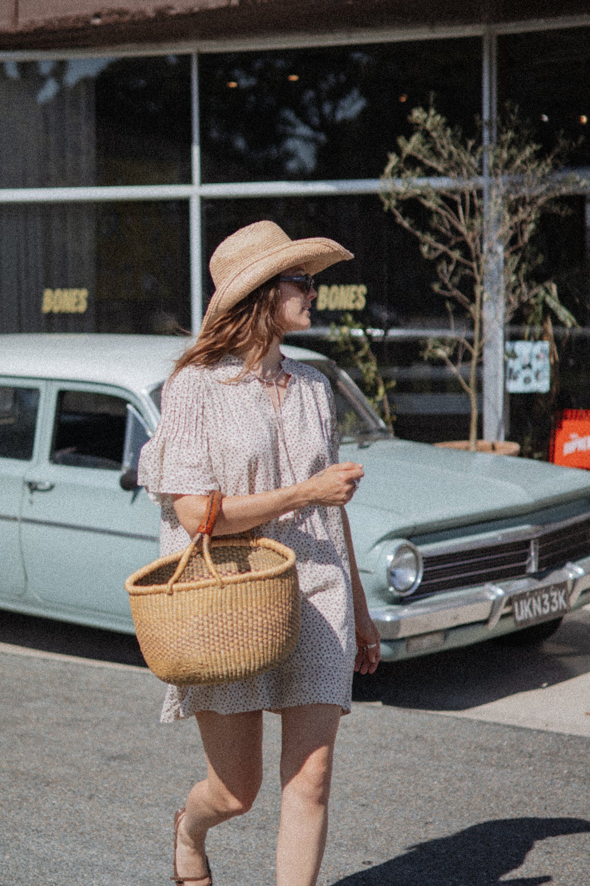 A woman walking outdoors in front of a vintage car and storefront, wearing a light beige patterned dress, wide-brim straw hat, and carrying a woven straw tote bag.