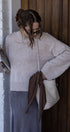 Woman in a light gray sweater and blue skirt holding a brown bag against a wooden wall.