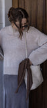 Woman in a light gray sweater and blue skirt holding a brown bag against a wooden wall.