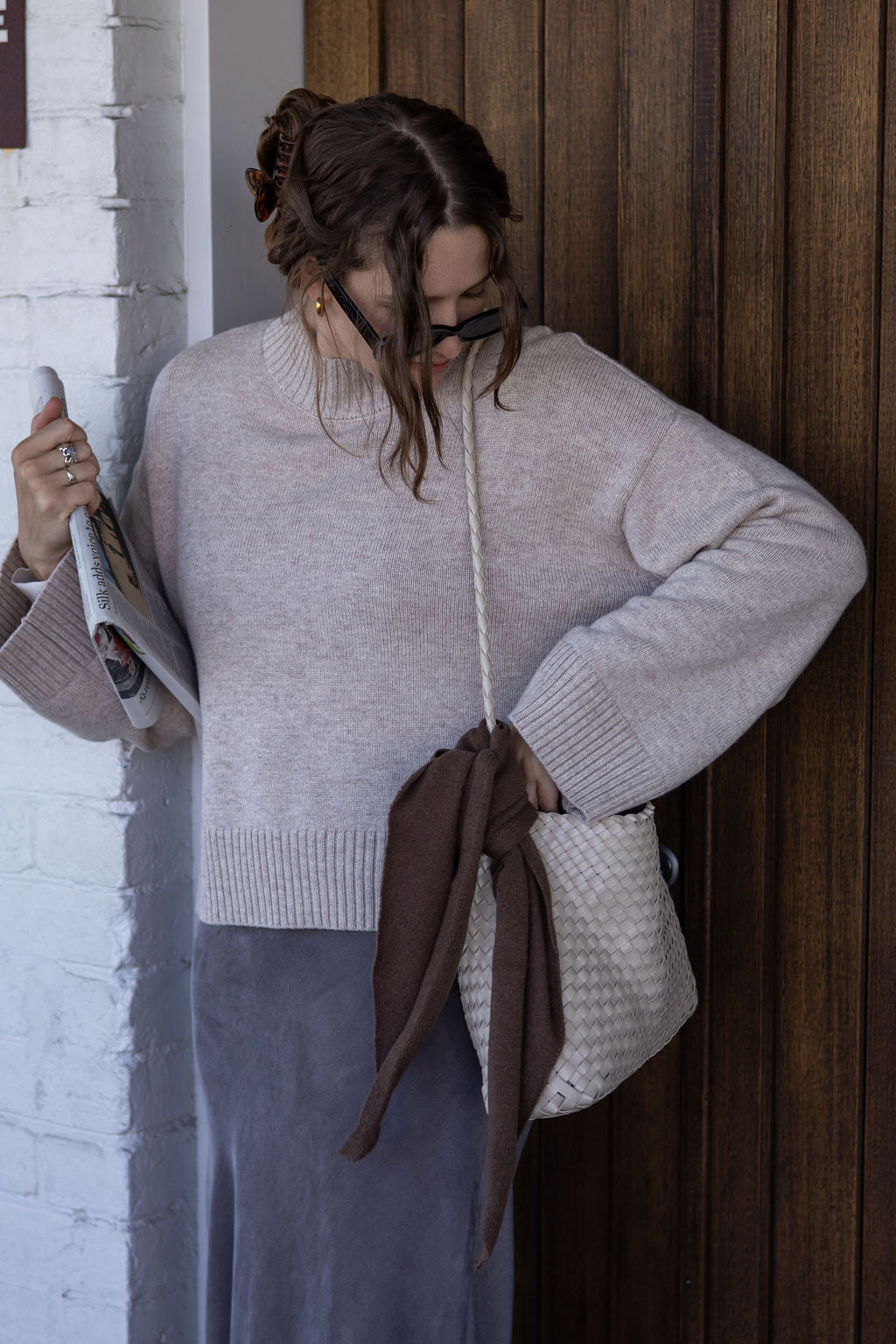 Woman in a light gray sweater and blue skirt holding a brown bag against a wooden wall.