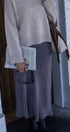 Person wearing a beige sweater and gray skirt holding a newspaper, standing against a white brick wall.
